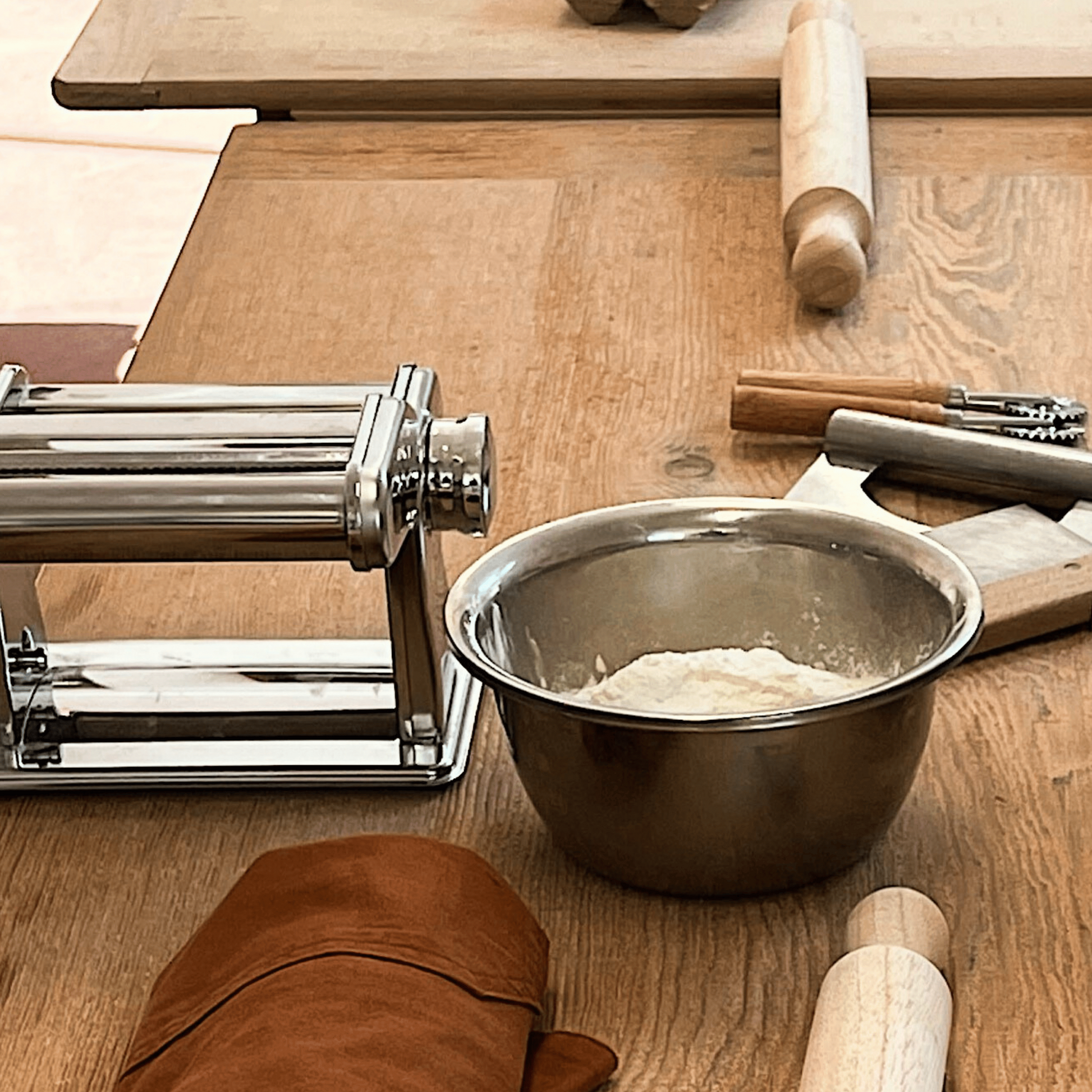 Pasta-making equipment including pasta machines, bowls of flour, and rolling pins on a wooden surface.